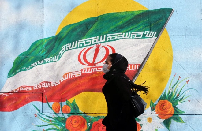 Một phụ nữ&nbsp;đeo khẩu trang đi bộ qua bức tường vẽ hình quốc kỳ Iran ở thủ đô Tehran.