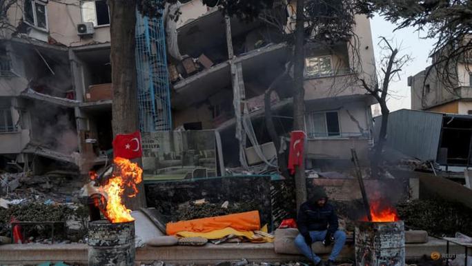 Một người đàn ông sưởi ấm bên đống lửa trước tòa nhà bị hư hại sau trận động đất ở Kahramanmaras hôm15-2. Ảnh: Reuters