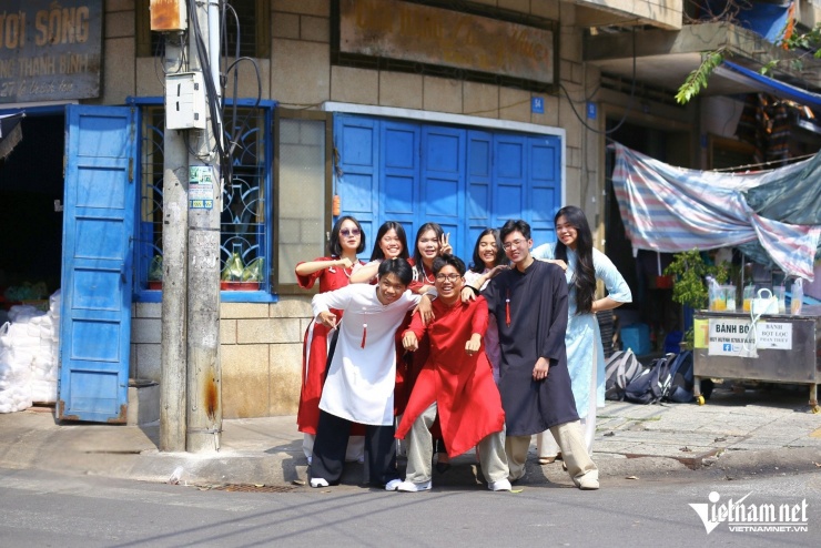 Nhóm bạn trẻ trong trang phục áo dài truyền thống và cách tân chụp ảnh lưu niệm trước dãy nhà cũ với cửa gỗ xanh đặc trưng của HTX Thanh Bình tại khu chợ