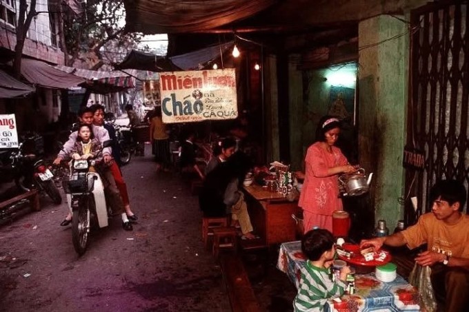 Người lái xe máy Honda len vào một quán ăn nhỏ trong ngõ. Quán bày bàn ghế đơn giản, thắp sáng bằng vài bóng đèn tròn, phía trên có tấm bạt nilon và biển hiệu viết tay. Hai bên là các ngôi nhà tập thể cũ, hình ảnh quen thuộc của phố phường Hà Nội thời trước.