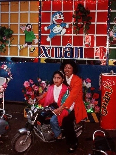 Người dân tạo dáng chụp ảnh Tết trên xe máy, trước không gian trang trí hoa giả. Phía sau là hình hai nhân vật Doraemon và Nobita trong bộ truyện "Doraemon" của Nhật Bản. Tác phẩm lần đầu xuất bản ở Việt Nam năm 1992, gắn bó với thiếu nhi nhiều thế hệ.