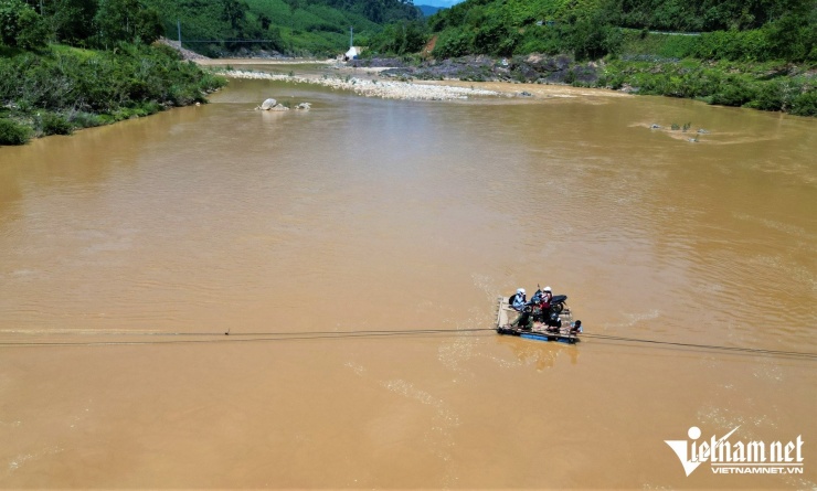 Chiếc bè tự chế chòng chành giữa sông, tiềm ẩn nguy cơ lật bất cứ lúc nào, trong khi cây cầu treo gần đó vẫn chưa hoàn thành.