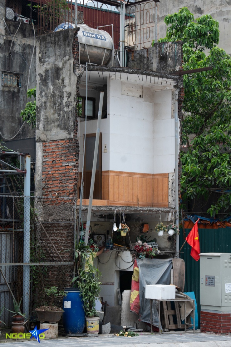 Căn nhà hai tầng trên phố Định Công sau giải tỏa hiện lên với hình hài méo mó, bị cắt xén gần như toàn bộ phần phía trước, chỉ còn trơ lại một lát cắt hẹp sâu vào bên trong. Những mảng tường gạch cũ lộ thiên, nham nhở, xen lẫn lớp bê tông bị đục bỏ dang dở, để lộ cả cốt thép han gỉ. Không gian bên trong bị thu hẹp tối đa, đủ đặt một cửa ra vào và phần tường ốp dở, phía trên vẫn còn bồn nước và lan can sắt cũ kỹ chênh vênh.