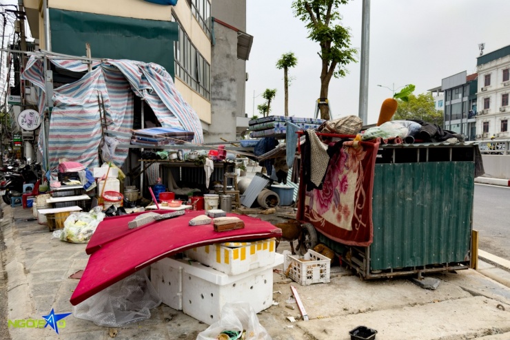 Cách đó vài trăm mét, tại một góc vỉa hè sát trục đường lớn, một hộ gia đình dựng lều tạm bằng bạt và vật liệu thô sơ để sinh hoạt. Không gian sống chắp vá từ thùng tôn, đệm, thùng xốp và các can nước, đồ dùng sinh hoạt xếp ngổn ngang; mọi hoạt động ăn uống, nghỉ ngơi đều diễn ra lộ thiên sát dòng xe cộ.