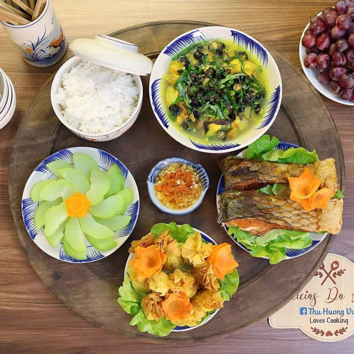 Mâm cơm gồm các món ốc om chuối đậu, cá chép sông rán giòn, chả giò nhân gà ngô non, su su luộc, tráng miệng bằng nho đỏ kẹo.