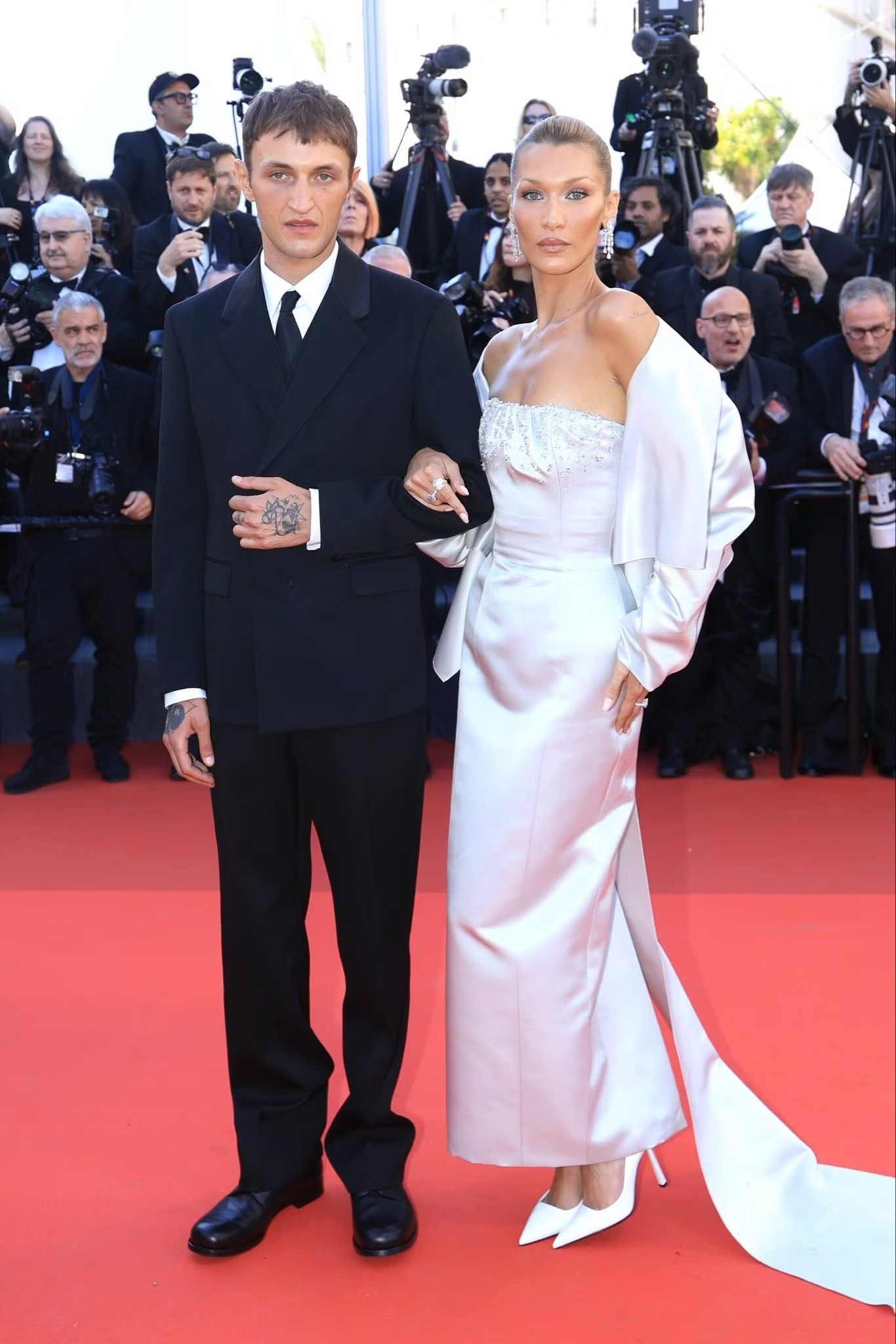 Siêu mẫu Bella Hadid chọn kiểu tóc búi cao kiểu diva cổ điển, làn da rám nắng nhẹ cùng bộ váy Prada may đo riêng. Thiết kế corset được đính pha lê ở phần cổ tim, kết hợp cùng trang sức Chopard. Bella tiếp tục chứng minh sức hút tại Cannes khi xuất hiện cùng em trai Anwar Hadid.