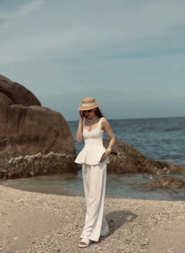Điểm nhấn của set đồ nằm ở cách phối phụ kiện đầy tinh tế. Mũ cói boater mang hơi thở cổ điển, kết hợp cùng chuỗi vòng cổ và khuyên tai ngọc trai tạo cảm giác sang trọng nhưng vẫn rất phù hợp với khung cảnh biển trời. Cô hoàn thiện outfit bằng sandal trắng và vòng tay bản lớn màu vàng ánh kim của Chanel có giá hơn 25 triệu, vừa đủ nổi bật nhưng không hề phô trương.
