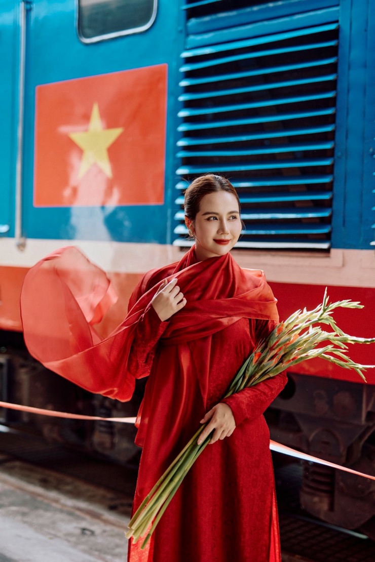 Cô chọn thiết kế áo dài đỏ kết hợp với khăn choàng đỏ, bối cảnh ga tàu có lá cờ Tổ quốc đầy ý nghĩa.