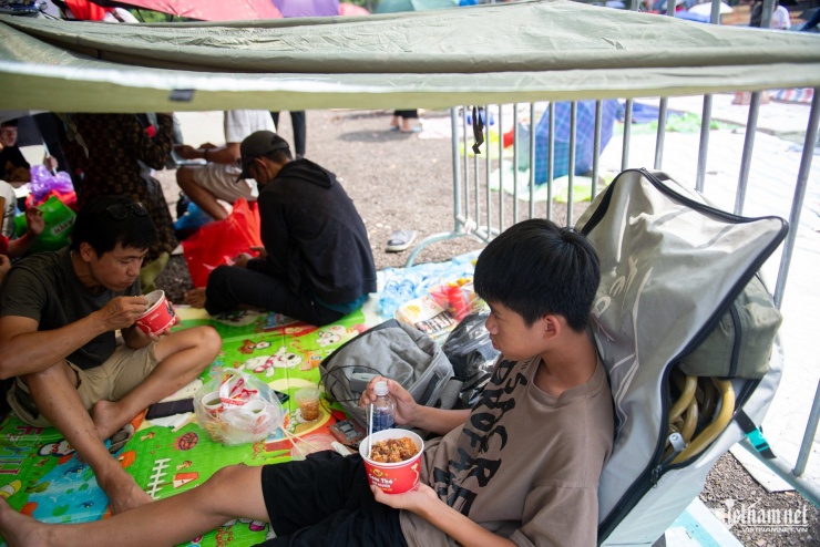 Khu vực nhà bạt chờ ở đây đã chật kín người từ sớm. Người dân tranh thủ ăn trưa và nằm nghỉ ngơi, nhiều gia đình mang sẵn đồ ăn, cũng có những người gọi dịch vụ giao hàng.
