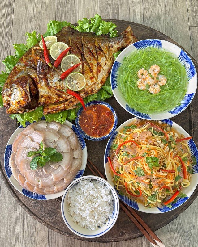 Không cần quá nhiều món, mâm cơm vẫn đủ vị với cá chim nướng vàng thơm, da giòn nhẹ và cay thơm vị muối ớt. Thịt ba chỉ luộc mềm ngọt ăn cùng mắm tôm chua đậm đà, trong khi nộm sứa xoài xanh đem lại vị giòn mát, chua ngọt. Bát canh bầu nấu tôm thanh nhẹ giúp cân bằng cả bữa cơm.