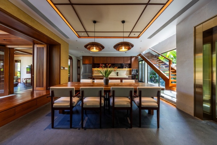 Bàn ăn dài đặt cạnh bếp, hướng ra mảng xanh bên ngoài. Khu bếp có hệ tủ áp tường và đảo bếp, giữ khoảng lưu thông ở giữa. Cửa mở và mảng kính cạnh cầu thang bổ sung ánh sáng tự nhiên cho khu vực phía trong.