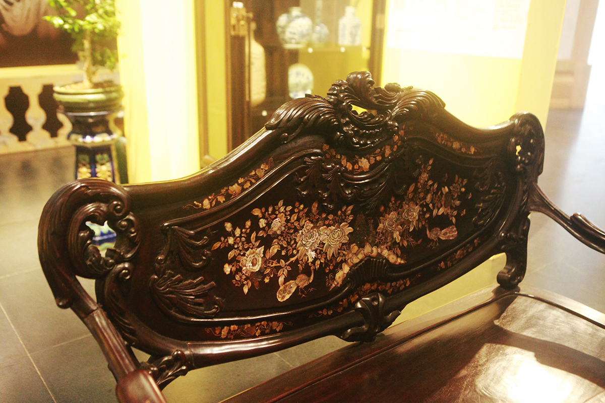 Bộ bàn ghế gỗ gụ, sơn nâu khảm trai ở không gian phòng khách được chạm lộng, chạm thủng đề tài hoa lá, chim, bướm, bầu rượu, túi thơ…