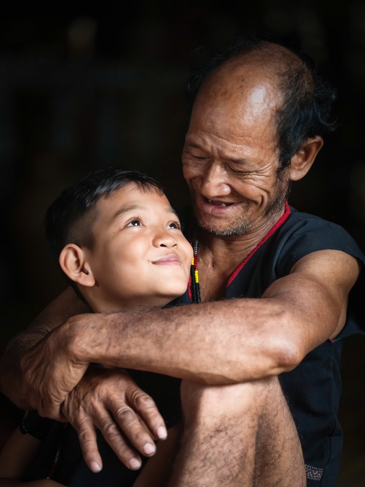 Trẻ nhỏ trong buôn có sự gắn bó sâu sắc với người lớn. Mối liên kết thiêng liêng ấy càng thêm bền chặt khi chúng được chính những người ông, người cha của mình kiên nhẫn truyền dạy cách làm nghề thủ công truyền thống. Ảnh: Ngô Trần Hải An. 
