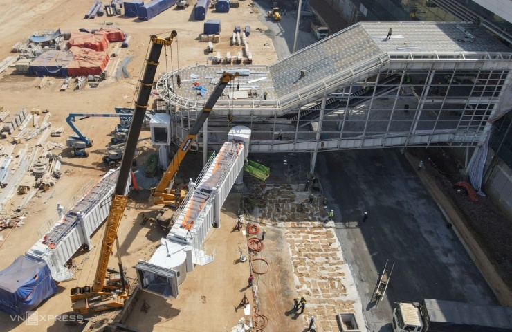 Xe cẩu lắp đặt cầu ống lồng, với hai cần cẩu 140 tấn phục vụ nâng hạ. Hàng trăm công nhân thi công khung, lợp mái, ốp kính các ống lồng còn lại. Theo thiết kế, sân bay Long Thành sẽ có 32 cầu ống lồng.