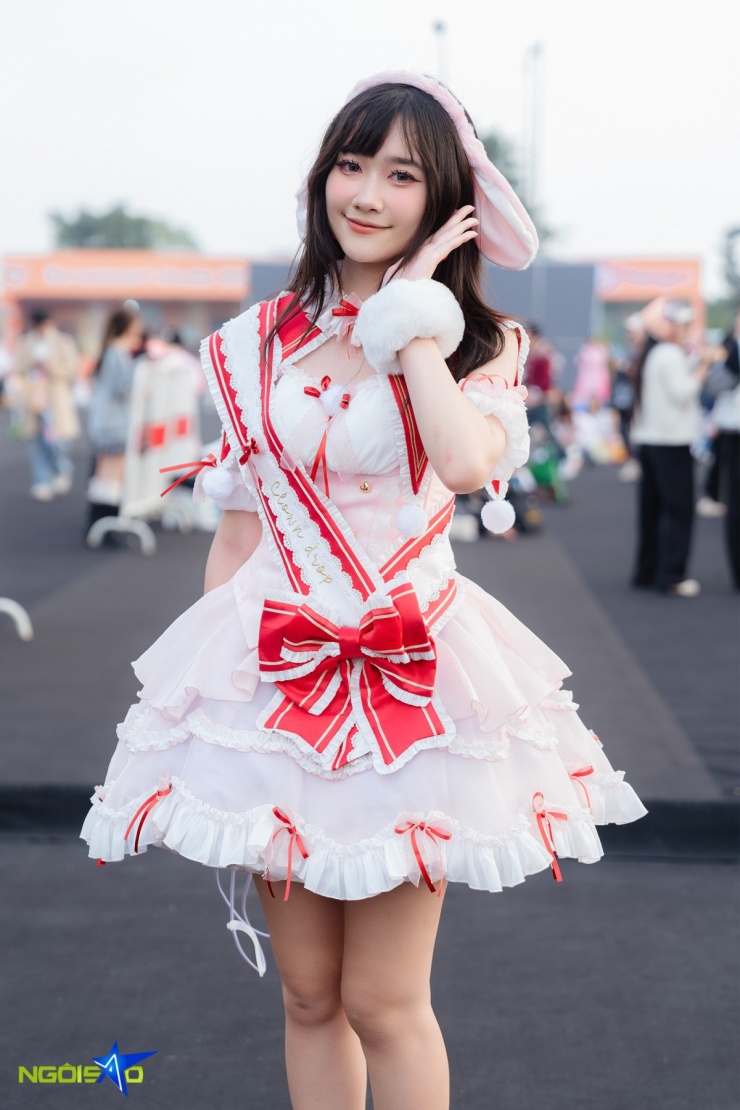 Nguyễn Trà My, 26 tuổi đến từ Hải Phòng, diện bộ váy phong cách dễ thương với tông trắng-hồng chủ đạo, có điểm nhấn là các dải ruy băng và nơ đỏ nổi bật. Thiết kế váy nhiều tầng với lớp voan và đường viền ren tạo vẻ bồng bềnh, nữ tính. Cô kết hợp thêm tai thỏ, cổ nơ và bao tay lông, giúp tổng thể trở nên trẻ trung và đáng yêu, phù hợp với không khí lễ hội.