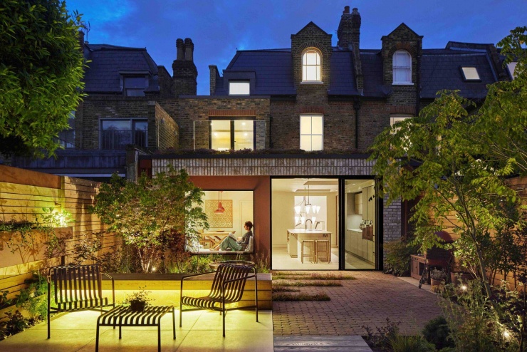 Toàn cảnh mặt sau ngôi nhà vào buổi tối, nơi ánh sáng vàng từ nội thất hắt ra khu vườn xanh mát. Thiết kế cửa kính lớn giúp ngôi nhà kết nối trực tiếp với không gian ngoài trời và tạo cảm giác ấm áp khi đêm xuống. Nguồn: Archello