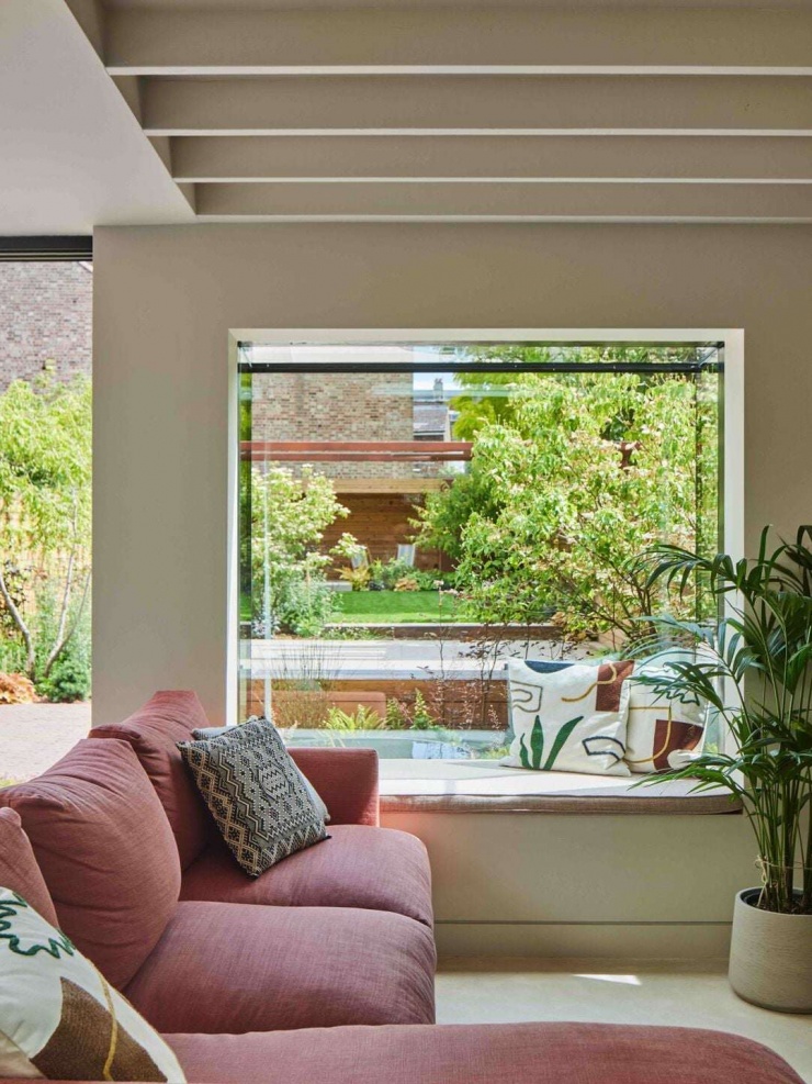 Phòng khách được bố trí tối giản với sofa màu trầm ấm và băng ghế cửa sổ nhìn ra vườn. Ô cửa lớn đóng vai trò như một “khung tranh xanh”, mang thiên nhiên trở thành một phần của nội thất. Nguồn: Archello