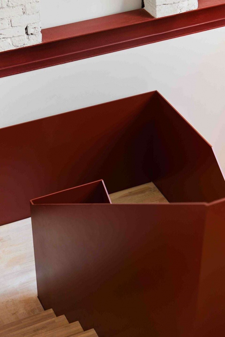 Chi tiết lan can và thân cầu thang thép gấp nếp tạo cảm giác liền khối, chắc chắn nhưng không nặng nề. Màu thép đỏ gỉ tương phản với tường trắng và gạch cũ, làm nổi bật tinh thần giao thoa giữa cũ và mới