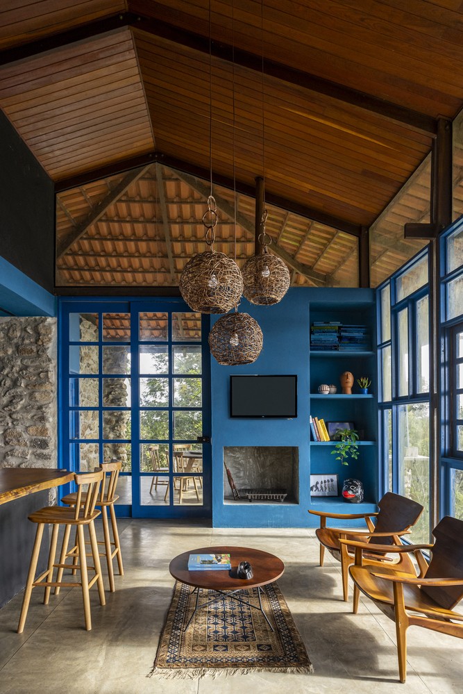 Bên trong căn nhà, trần gỗ mái dốc kết hợp hệ đèn tre thủ công tạo cảm giác ấm áp cho không gian sinh hoạt chung. Tường sơn xanh lam cùng hệ cửa kính và cửa đồng bộ màu sắc.