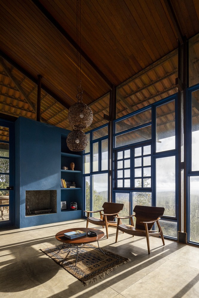 Không gian phòng khách mở hoàn toàn ra thiên nhiên. Nội thất gọn nhẹ với ghế gỗ, bàn tròn và thảm dệt tay đặt trên nền xi măng mài.