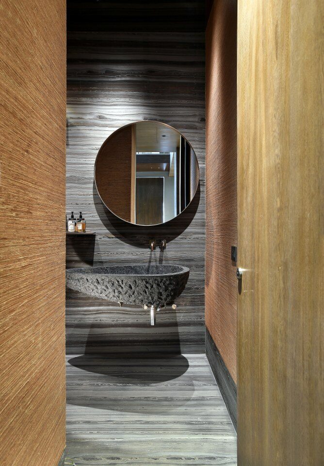 Khu vực lavabo sử dụng đá xám ốp xuyên suốt tường và sàn, kết hợp bồn rửa bằng đá có bề mặt gia công thô. Gương tròn treo tường, vòi nước âm tường và hệ cửa gỗ giúp tối giản chi tiết.
